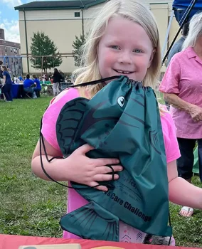 A young girl with long blonde hair smiles widely while holding a green drawstring bag featuring a fish print. She wears a pink t-shirt, and a lively outdoor event can be seen in the background.