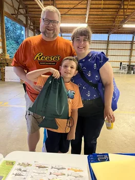 A smiling family of three poses for a photo indoors. The father, wearing an orange Reese's shirt, stands on the left with his arm around his son, who is holding a green drawstring bag. The mother, in a blue top with a pattern, stands on the right. A table with informational materials is visible in the foreground.