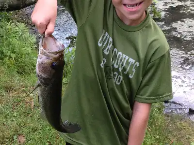 A smiling child in a green Cub Scouts shirt holds up a large fish by its mouth, standing near a grassy area next to a body of water.