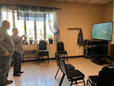 Two people stand indoors, one holding a fishing rod aimed at a TV displaying a fish image. Chairs and potted plants by a window set a casual tone.