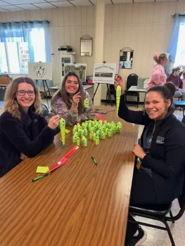 Three smiling people seated at a table display bright green fish lures. The room is decorated with motivational posters and has a lively, cheerful atmosphere.