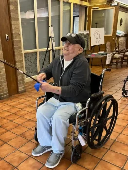 A smiling man in a wheelchair holds a fishing rod inside a community center. He wears a cap, glasses, and casual attire. The atmosphere is relaxed and joyful.