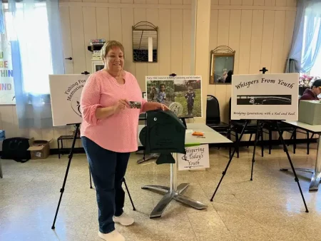A smiling woman in a pink sweater and jeans holds a card. Behind her are posters with text “Fishing for Memories” and “Whispers From Youth.”