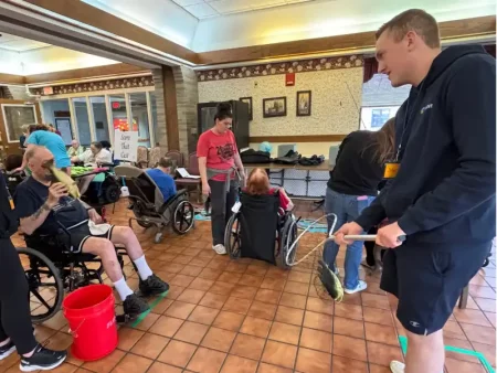 A lively scene in a community center shows people of various ages, some in wheelchairs, engaging in activities. One man in a wheelchair holds fish props by a red bucket, while another person stands nearby with a net. The atmosphere is active and social.